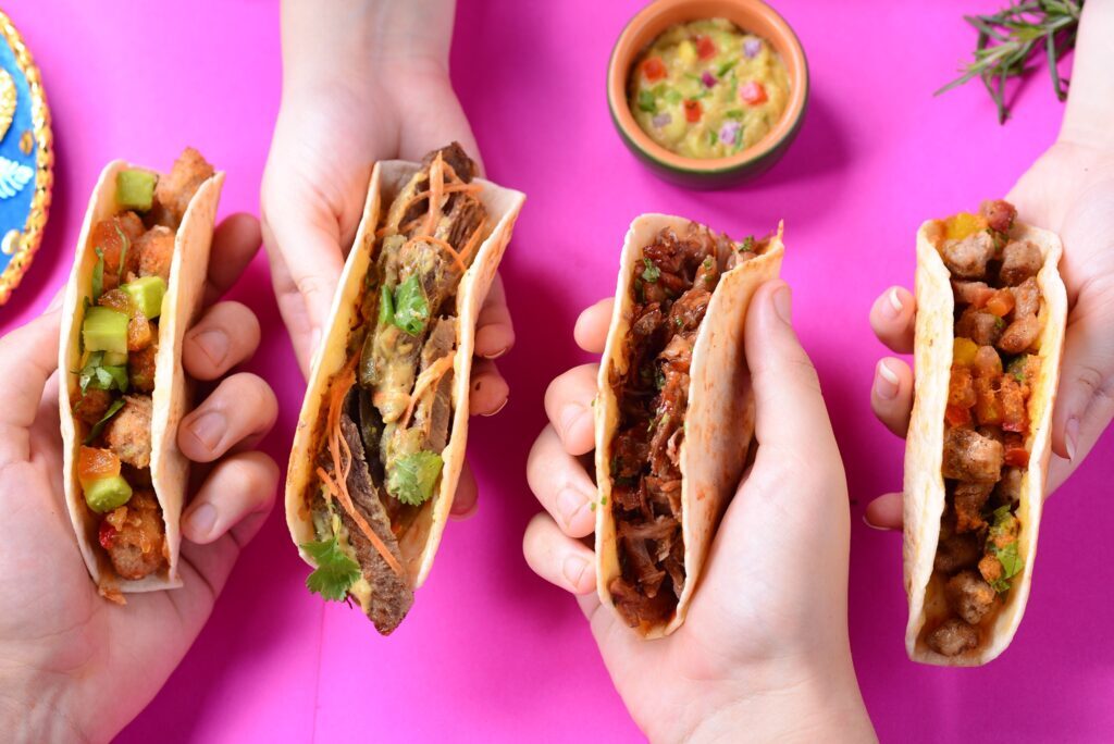 Imagem com mãos segurando tacos de diferentes sabores Guacamole Porto Alegre.